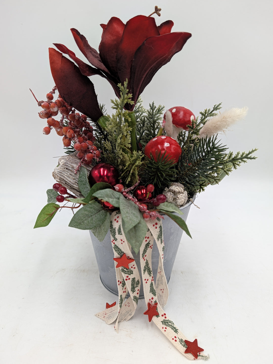 Weihnachtsgesteck im Zinktopf mit dunkelroter Amaryllis, Tannenzweigen, Fliegenpilzen und Bändern mit roten Sternen