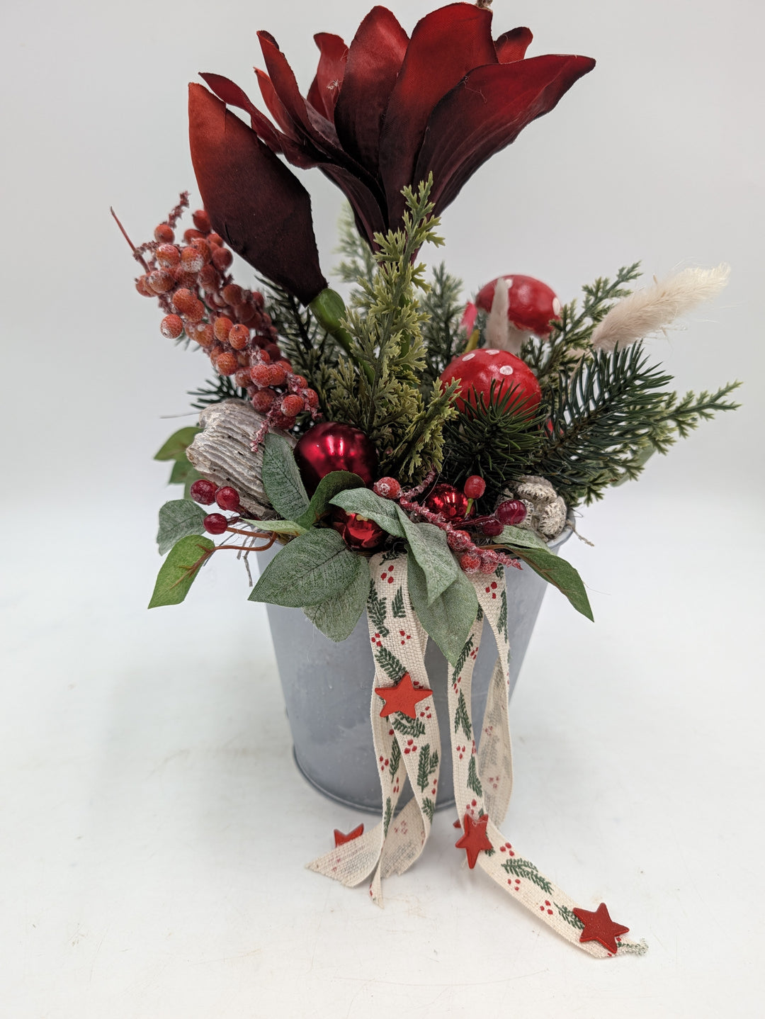 Seitenansicht des Gestecks: rote Amaryllis, Beeren mit Raureif, rote Kugel, Holzstück und Rispenhalm im Zinktopf