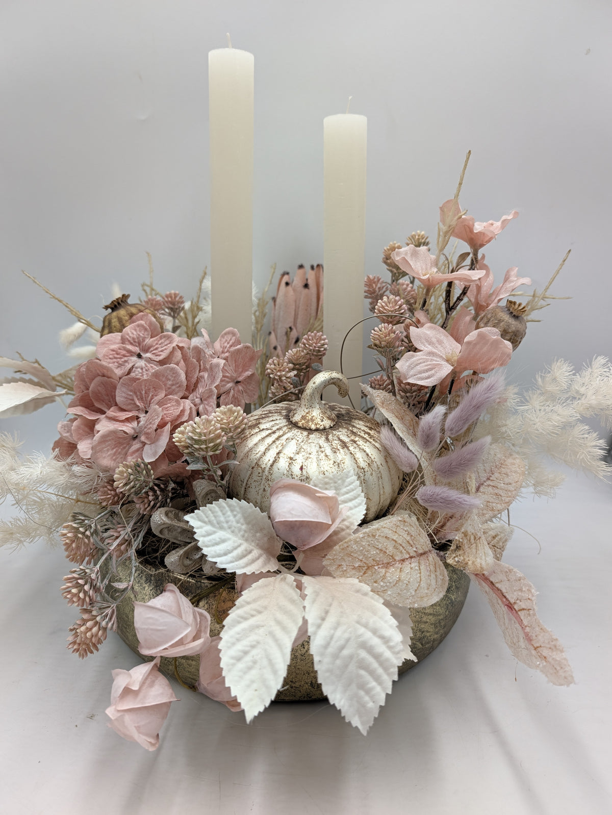 Herbstgesteck mit weißem Kürbis, rosa Hortensien, Rosenknospen und zwei cremefarbenen Stabkerzen in goldener Schale