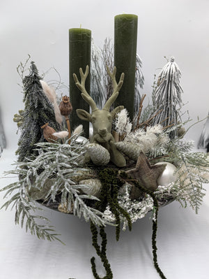 Wintergesteck mit grüner Hirschfigur, zwei olivgrünen Kerzen, beschneiten Zweigen und Zapfen in flacher Schale
