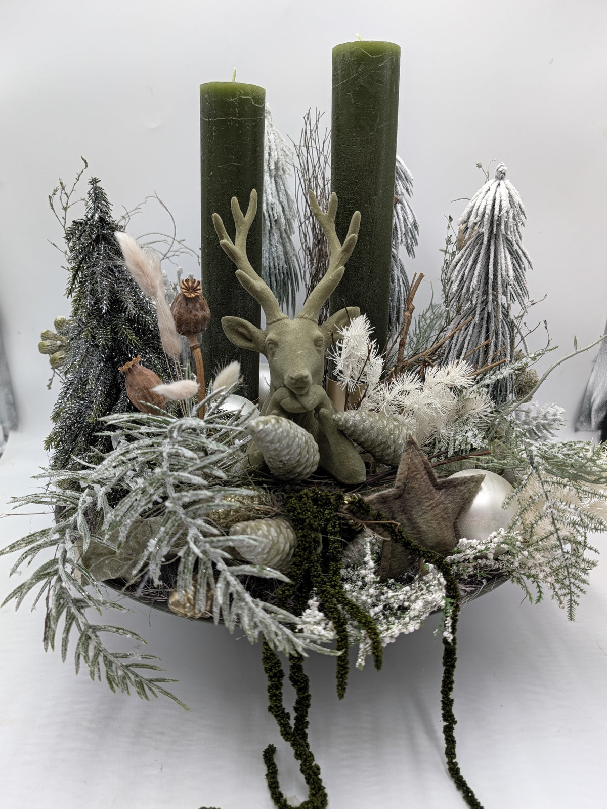 Wintergesteck mit grüner Hirschfigur, zwei olivgrünen Kerzen, beschneiten Zweigen und Zapfen in flacher Schale