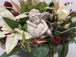Nahaufnahme des Engels mit Herz und Aufschrift In lieber Erinnerung zwischen weißen Lilien und Zapfen