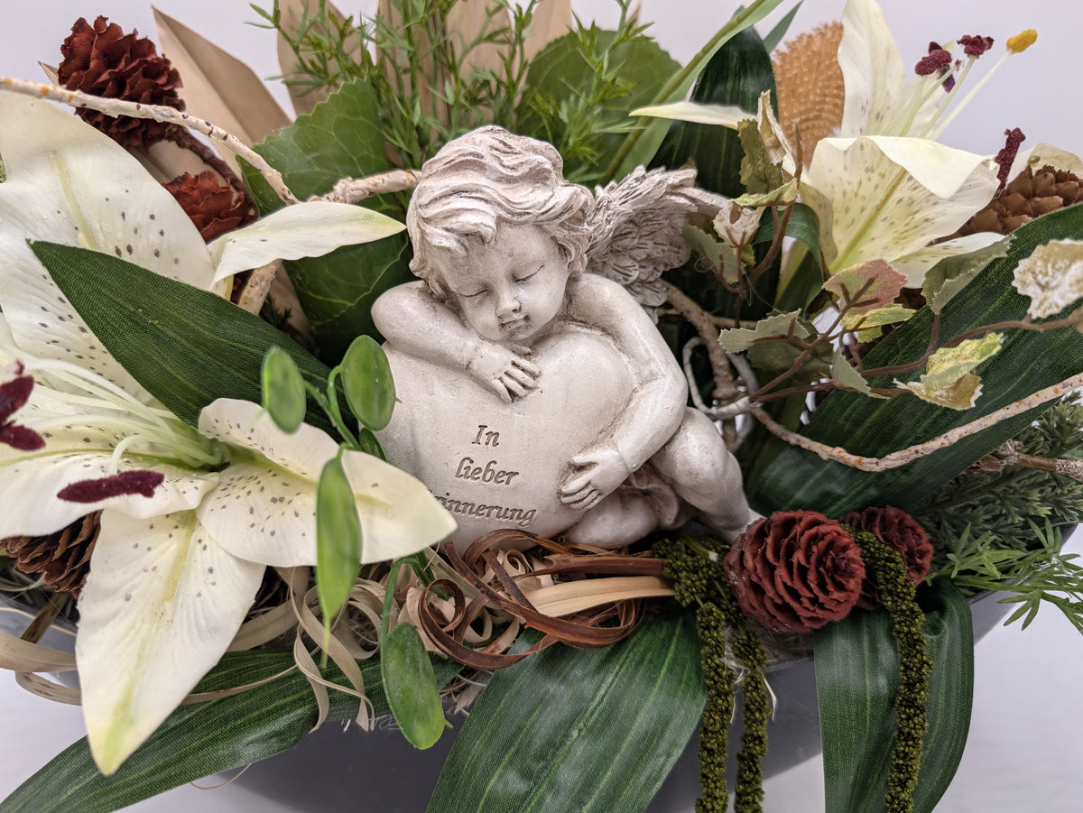 Nahaufnahme des Engels mit Herz und Aufschrift In lieber Erinnerung zwischen weißen Lilien und Zapfen