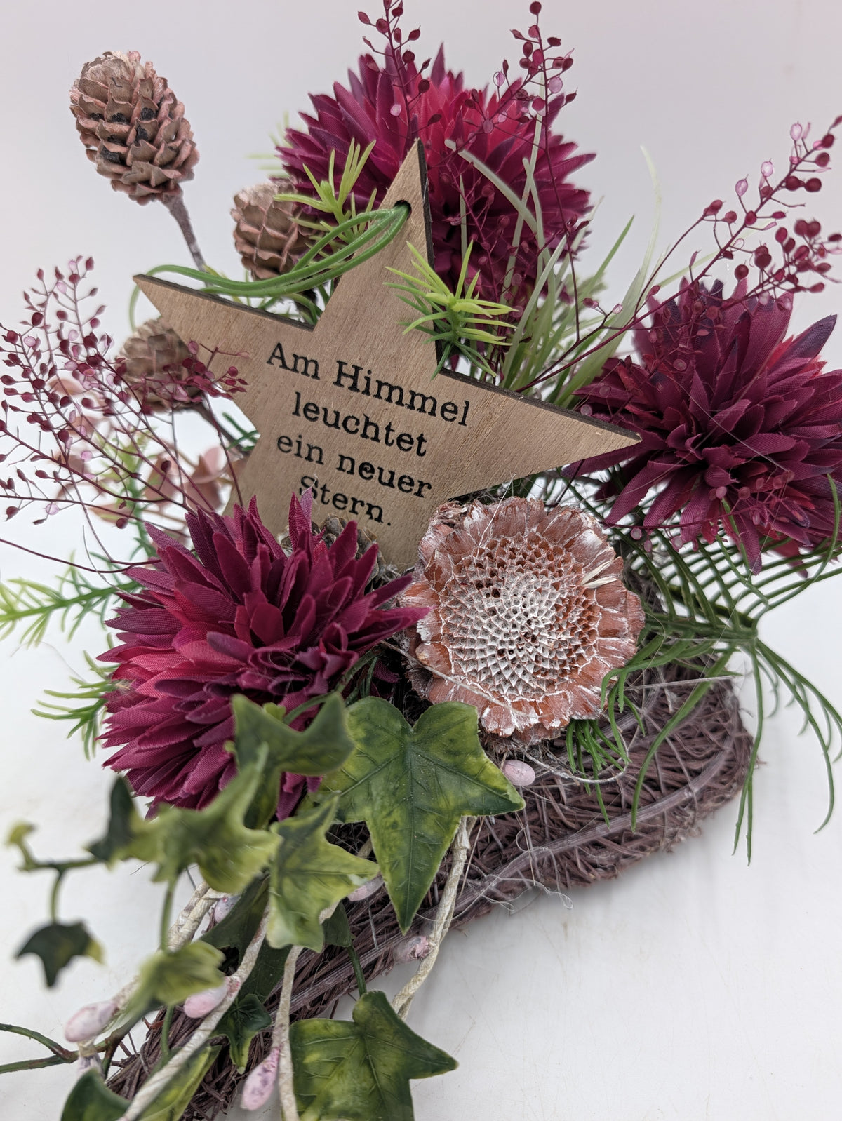 Holzstern mit Spruch zwischen beerenlila Dahlien, getrockneter Blüte, Zapfen und Gräsern auf einem Herz aus Reisig