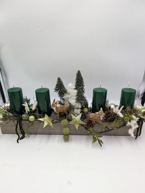 Langes Adventsgesteck von vorn: grauer Holzkasten, grüne Stumpenkerzen, Rehe und weiße Gräser über dem Rand