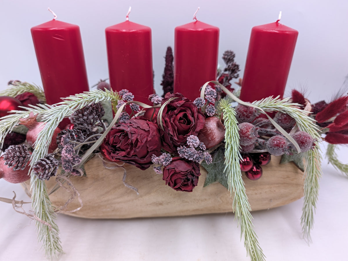 Nahaufnahme des Adventsgestecks mit dunkelroten Rosen, gefrosteten Beeren, Zapfen und roten Kugeln in heller Holzschale