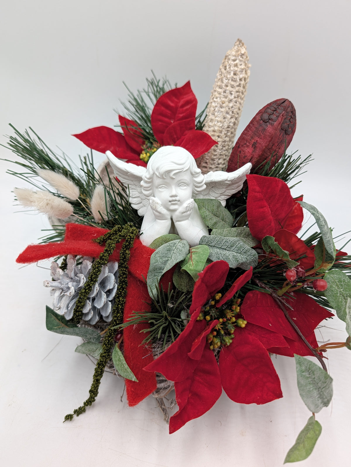 Weihnachtliches Gesteck im Korb mit Engel, roten Samtblüten, grau gekalkten Zapfen und roten Beeren