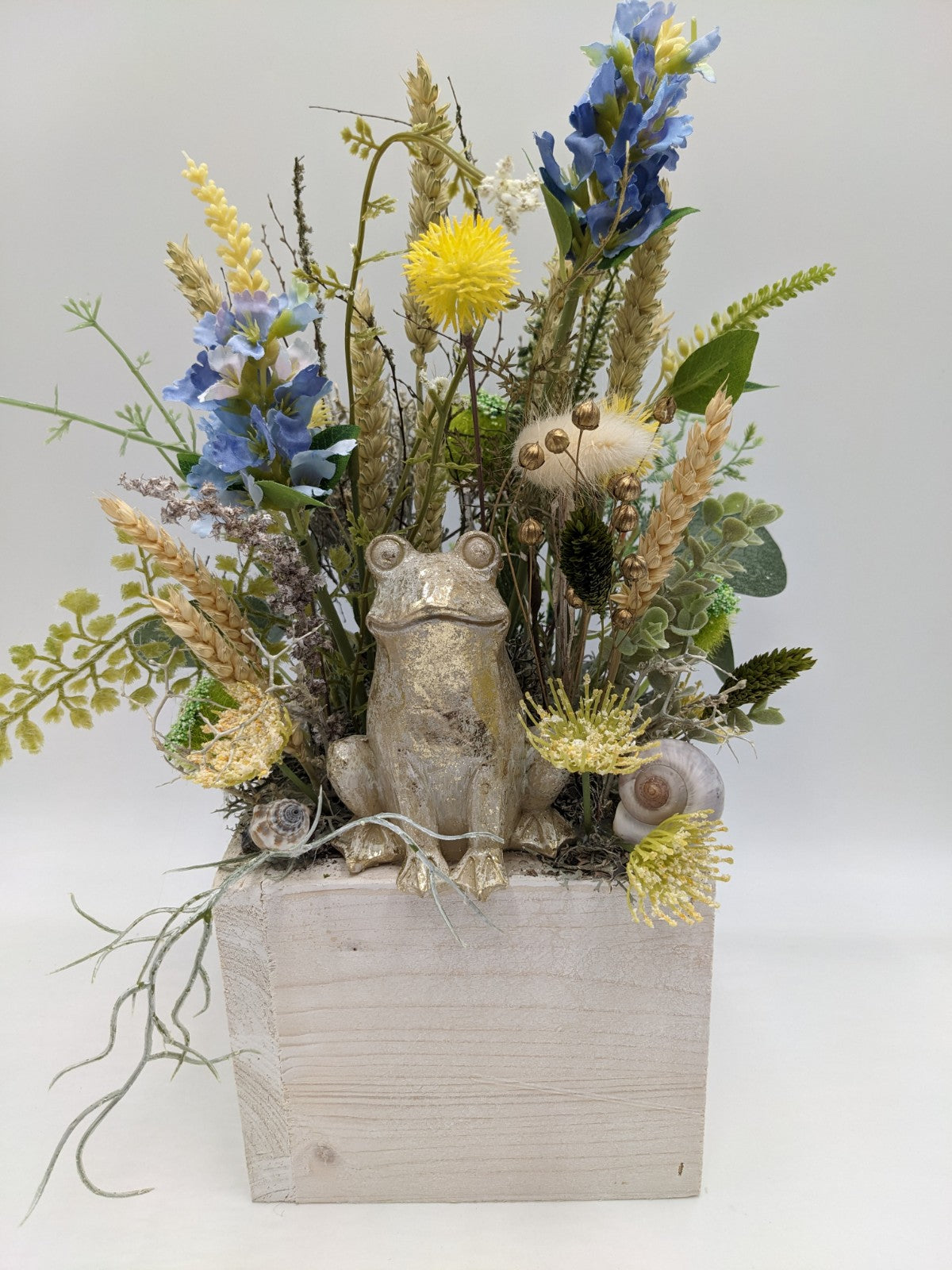 Sommergesteck in heller Holzlade mit goldenem Frosch, blauen Blüten, gelben Blütenköpfen und Ähren