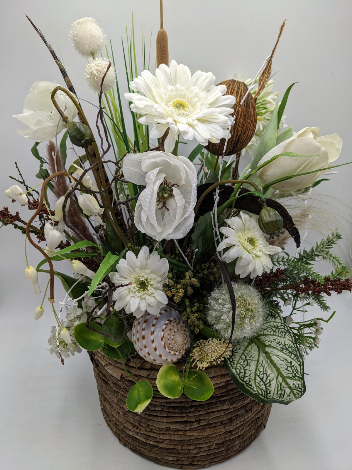 Nahansicht des Gestecks: weiße Gerbera, Mohnblüte, Rohrkolben und gepunktete Schneckenmuschel über dem Korbrand