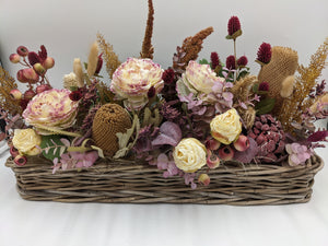 Langes Herbstgesteck im Weidenkorb mit cremefarbenen Rosen, weinroten Disteln, rosa Beeren und ockerfarbenen Gräsern