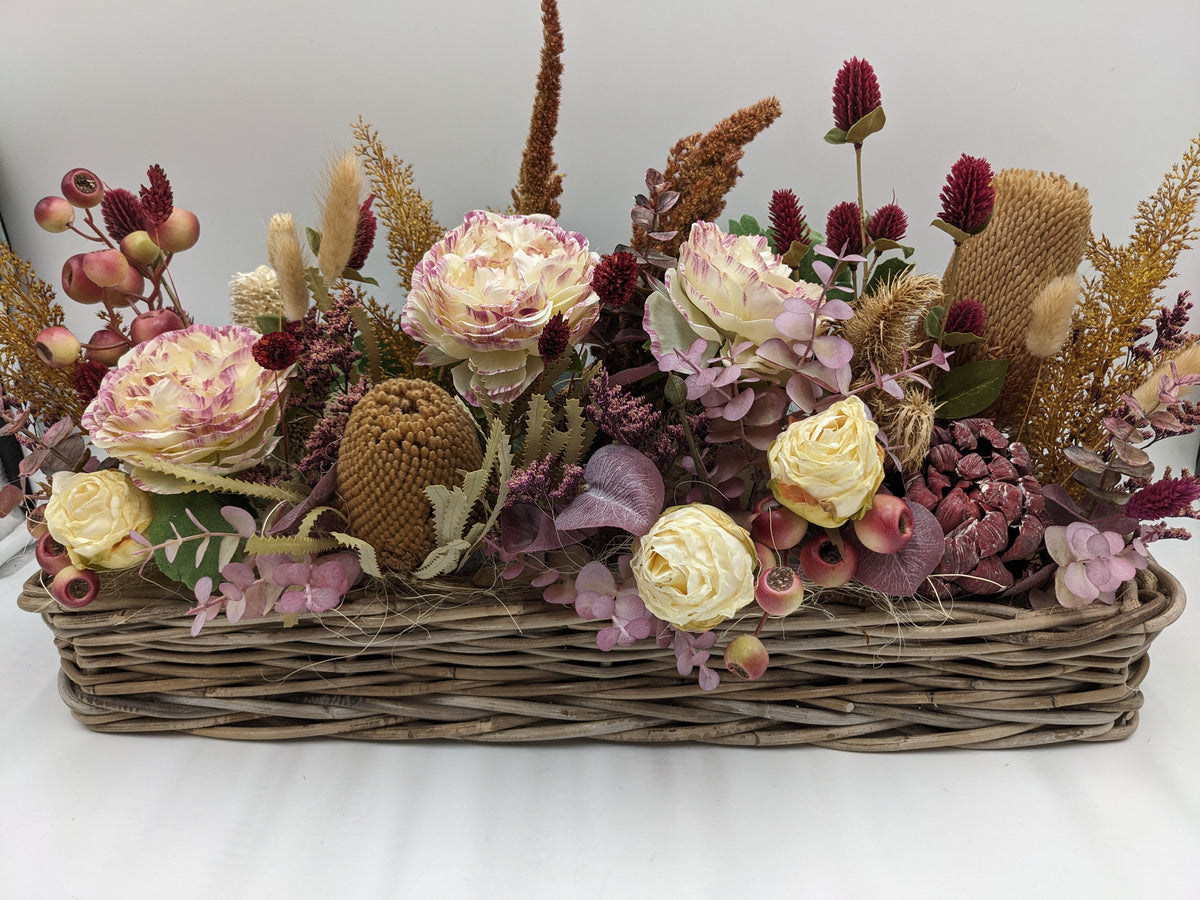 Langes Herbstgesteck im Weidenkorb mit cremefarbenen Rosen, weinroten Disteln, rosa Beeren und ockerfarbenen Gräsern
