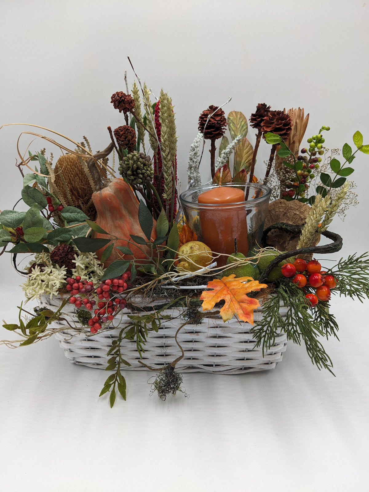 Herbstgesteck im weißen Korb mit Glaswindlicht, orangefarbener Kerze, Kürbis, Zapfen, roten Beeren und Gräsern
