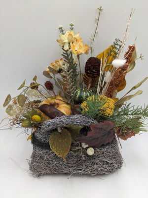 Herbstgesteck im Geästkorb mit orangefarbenem Kürbis, grünem Zierkürbis, Zapfen, cremefarbenen Blüten und Gräsern