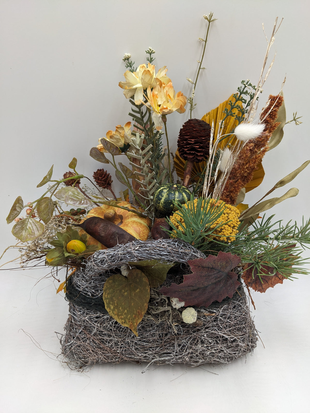 Herbstgesteck im Geästkorb mit orangefarbenem Kürbis, grünem Zierkürbis, Zapfen, cremefarbenen Blüten und Gräsern