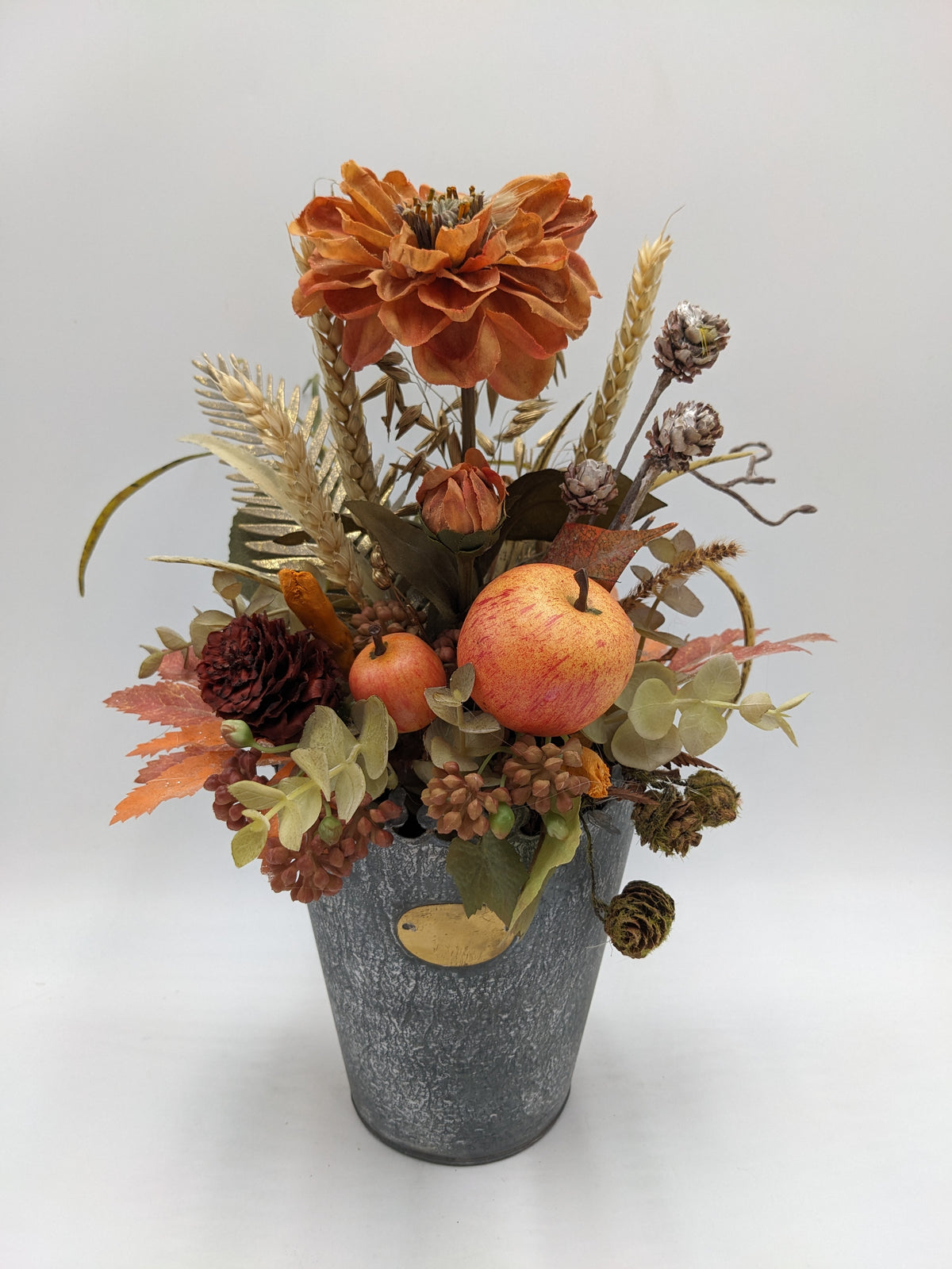 Herbstgesteck im grauen Zinktopf mit oranger Blüte, Äpfeln, Weizenähren, Zapfen und Herbstblättern