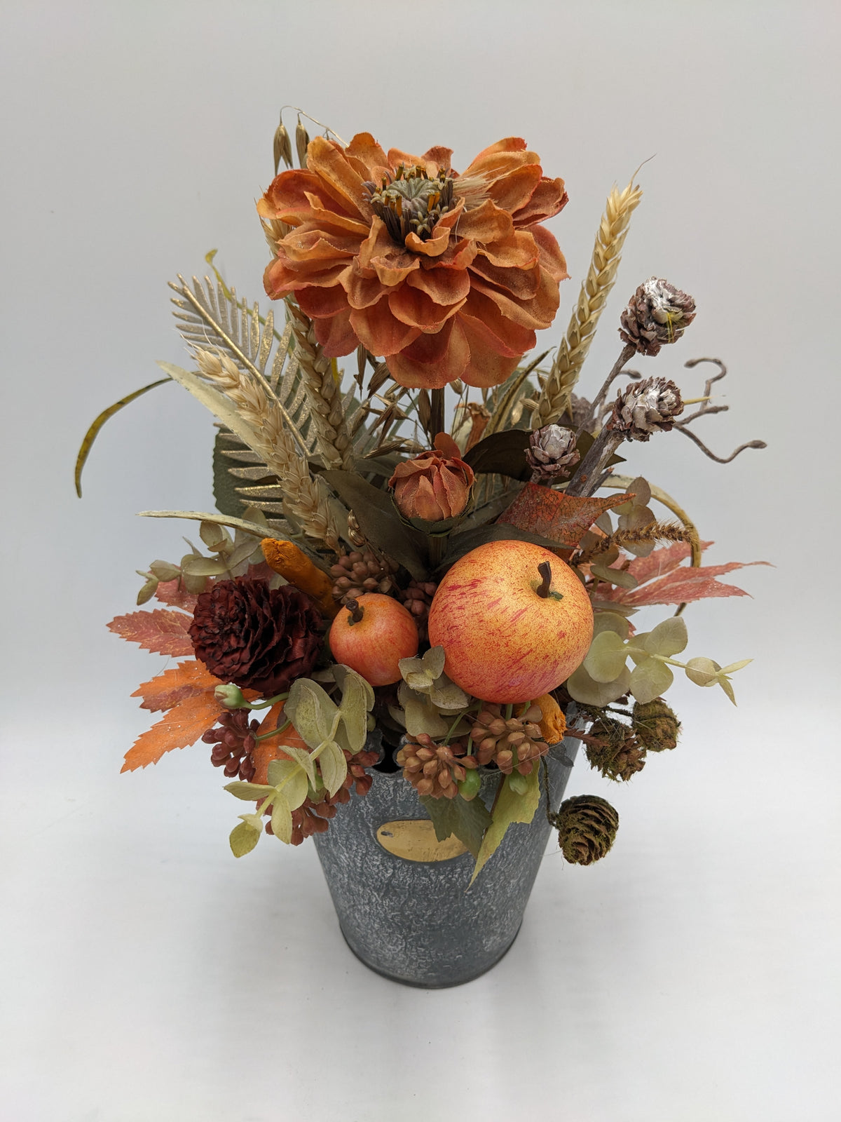 Herbstgesteck von oben: orange Blüte, zwei Äpfel, dunkelrote Blüte, Eukalyptus und Weizen im Zinktopf
