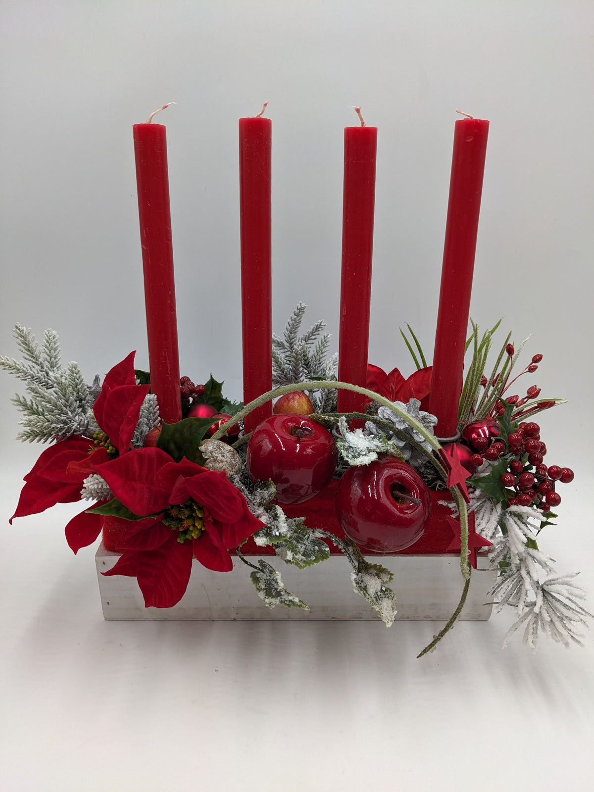 Adventsgesteck im weißen Holzkasten mit vier roten Stabkerzen, roten Äpfeln, Weihnachtssternen und Beeren