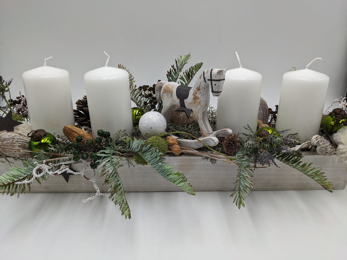 Langes Adventsgesteck im weißen Holztablett mit vier weißen Kerzen, Schaukelpferd, grünen Kugeln und Zapfen