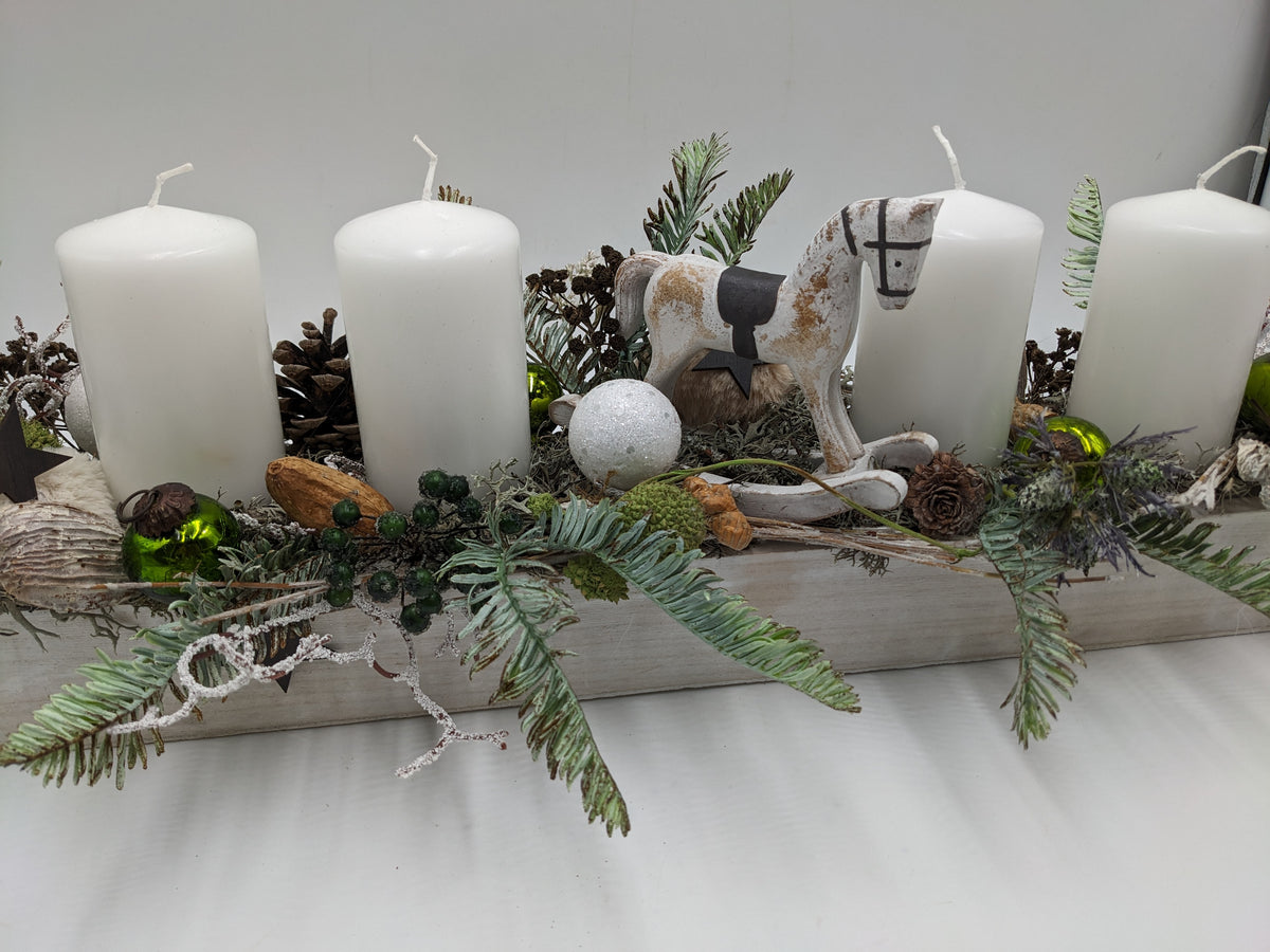 Seitenansicht des Adventsgestecks: Schaukelpferd aus Holz zwischen weißen Stumpenkerzen, Tanne und Beeren