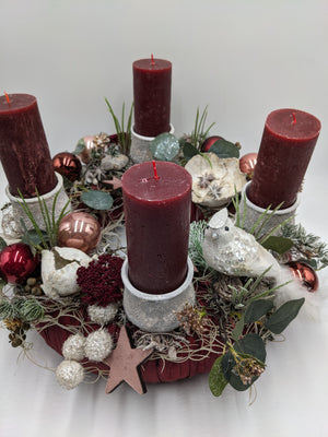 Adventsgesteck von schräg oben: dunkelroter Stoffring, vier rote Kerzen, Zapfen, rosa Holzstern, beschneite Zweige