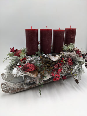 Holzschlitten von der Seite, darauf vier rote Kerzen, beschneite Tannenzweige, rote Beeren und Glitzersterne
