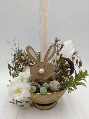 Nahaufnahme des Ostergestecks: Holzhase, gesprenkelte Eier, grüne Zuckerbeeren und weiße Blüten in goldener Fußschale