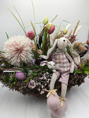 Stoffhase in kariertem Kittel sitzt auf dem Nestrand, dahinter rosa Tulpen, Ähren, Gräser und violette Ostereier