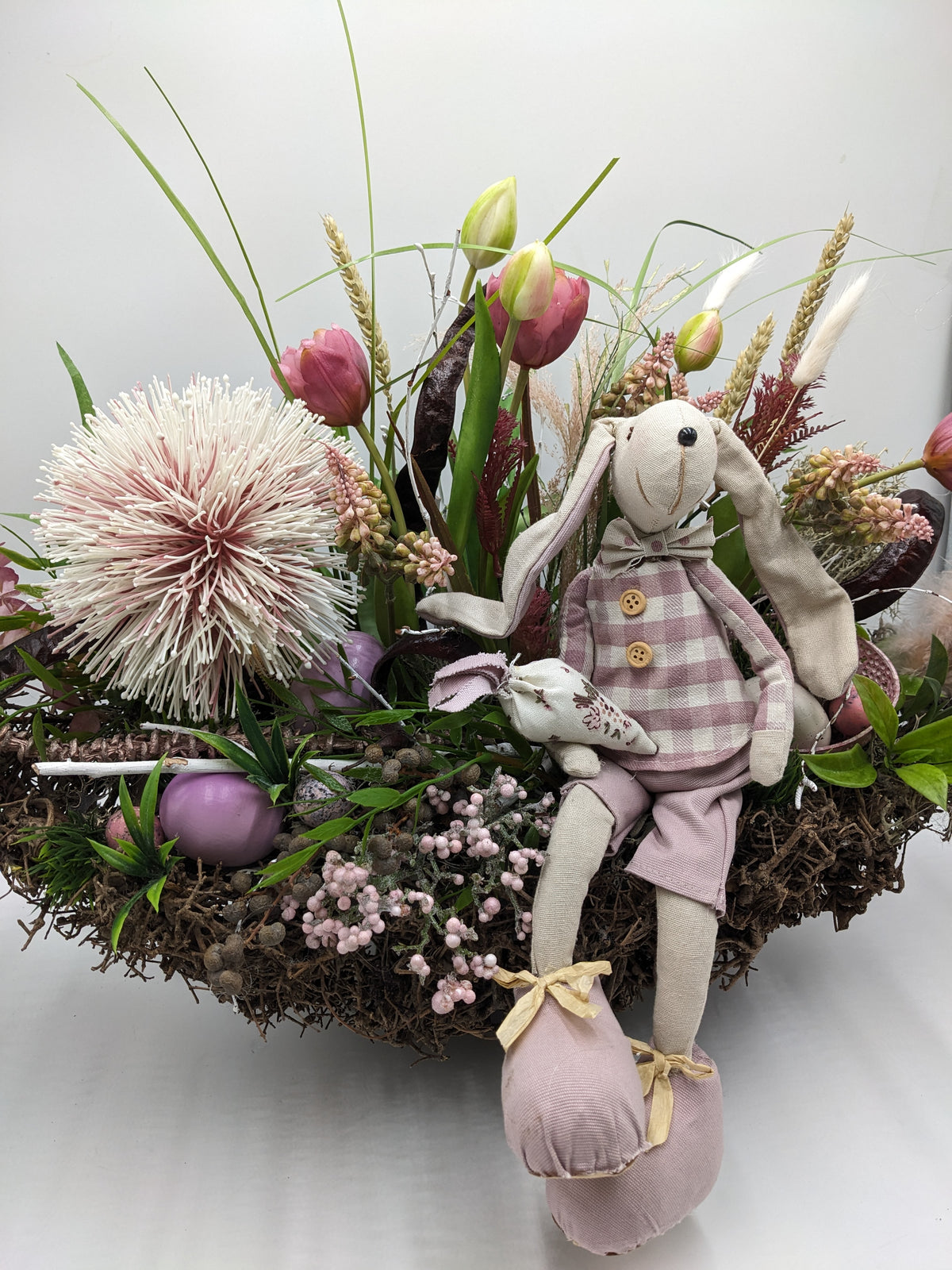Stoffhase in kariertem Kittel sitzt auf dem Nestrand, dahinter rosa Tulpen, Ähren, Gräser und violette Ostereier