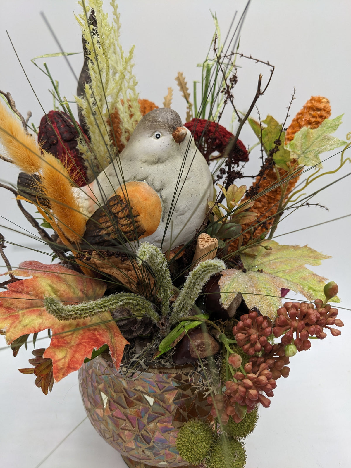Nahaufnahme: grau-weißer Keramikvogel mit oranger Brust zwischen Gräsern, Zapfen, Herbstlaub und roten Blüten