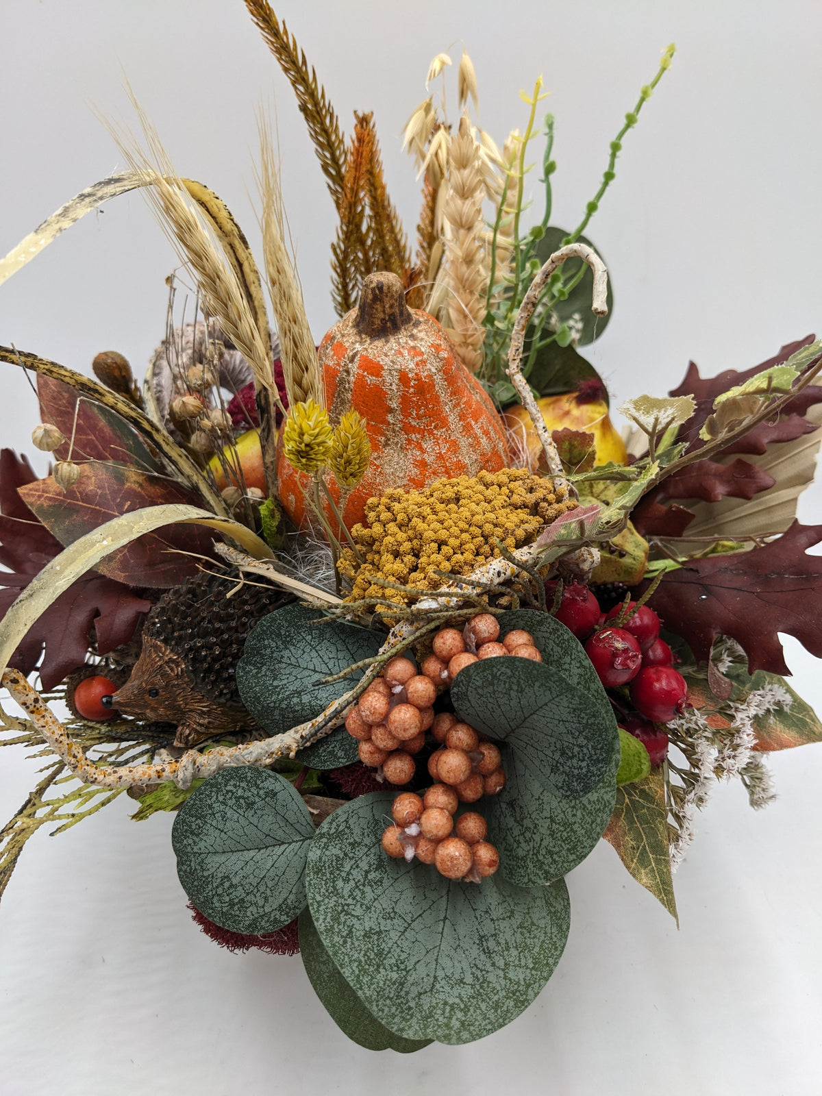 Nahaufnahme des Herbstgestecks: Kürbis, senfgelbe Dolden, apricotfarbene Beerentraube und dunkelrote Eichenblätter