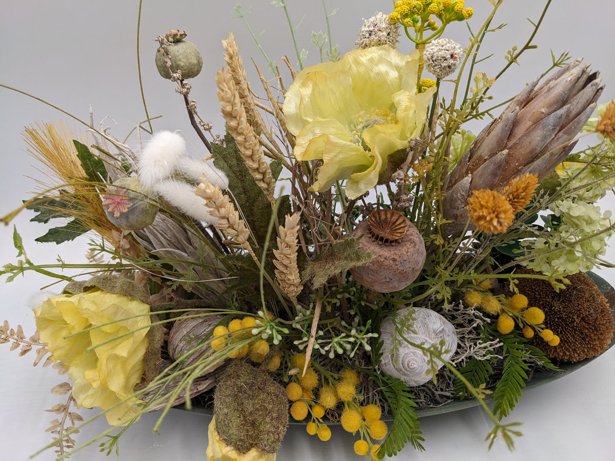 Nahaufnahme des Gestecks: gelbe Mohnblüte, braune Mohnkapsel, Protea, Farn und weißes Schneckenhaus im Moos