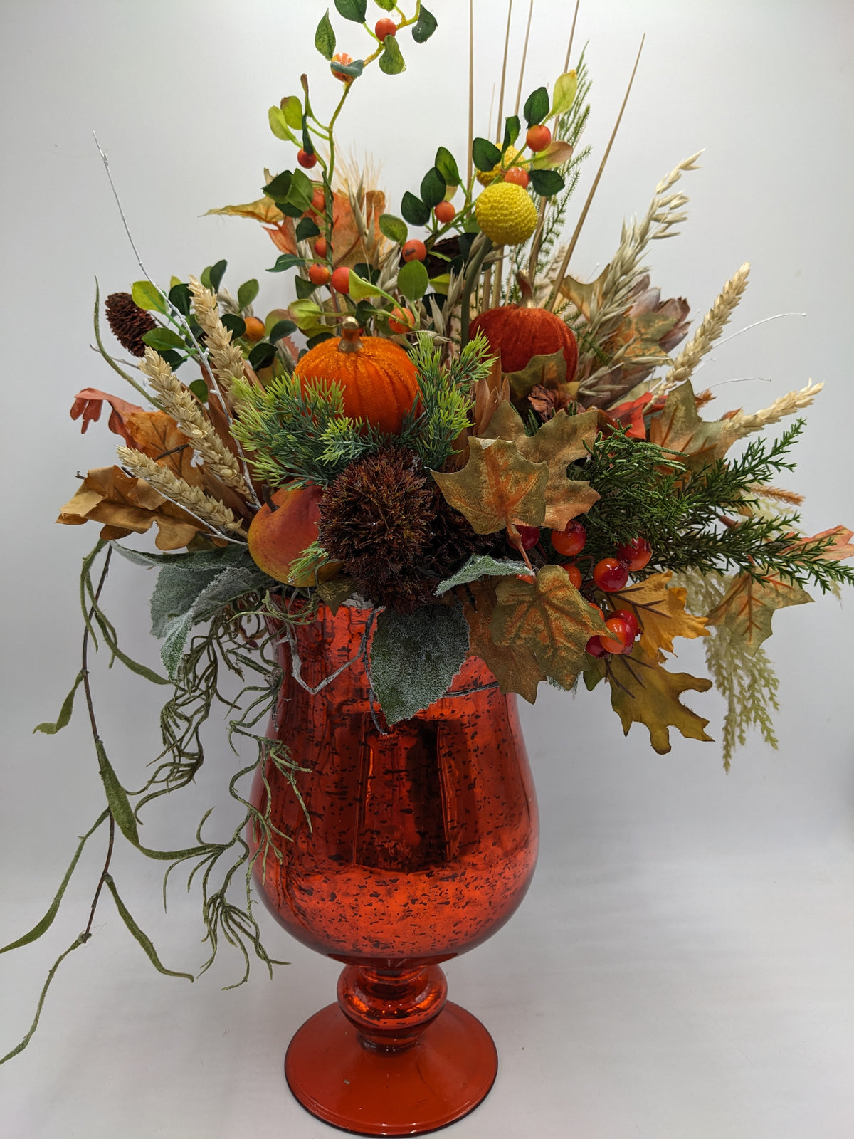Herbstgesteck im orangefarbenen Glaspokal mit Kürbissen, Beeren, Ahornblättern und hellen Weizenähren