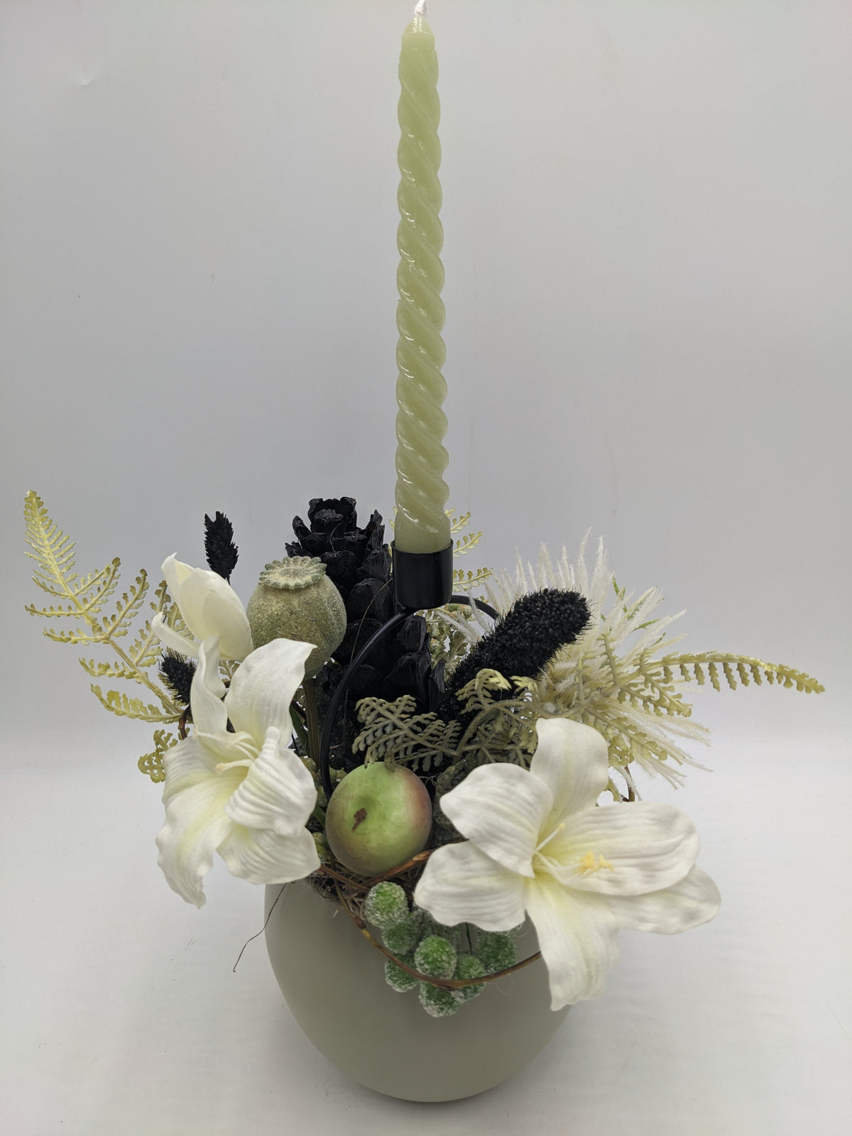 Herbstgesteck von vorn: weiße Amaryllis, grüner Apfel, schwarze Ähren, helle Farnwedel und Kerze im schwarzen Halter