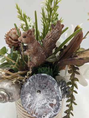Nahaufnahme der glitzernden Eichhörnchen-Figur zwischen Zapfen, weißen Blüten, Holzscheibe und Eukalyptusblättern