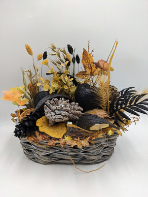 Herbstgesteck im grauen Weidenkorb mit schwarzen Glitzerfrüchten, hellen Zapfen, goldgelben Ähren und orangem Laub