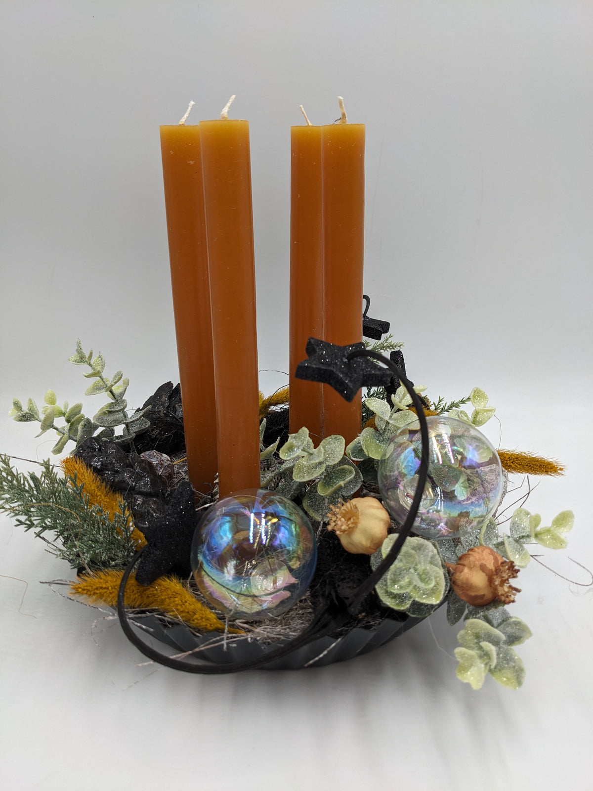 Adventsgesteck mit vier ockerfarbenen Stabkerzen, schillernden Glaskugeln, schwarzen Sternen und Eukalyptus