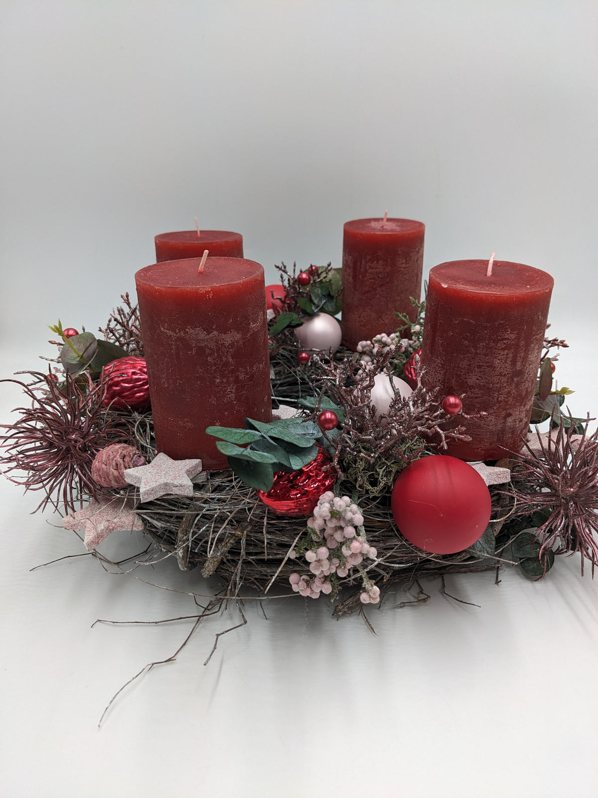 Adventsgesteck von der Seite: vier rote Stumpenkerzen auf Reisigkranz mit roten und rosa Kugeln, Sternen und Eukalyptus