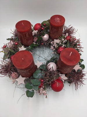 Adventsgesteck von oben: vier rote Kerzen im Kreis, rosa Glitzersterne, Beerenrispen, Zapfen und rötliche Spider-Blüten