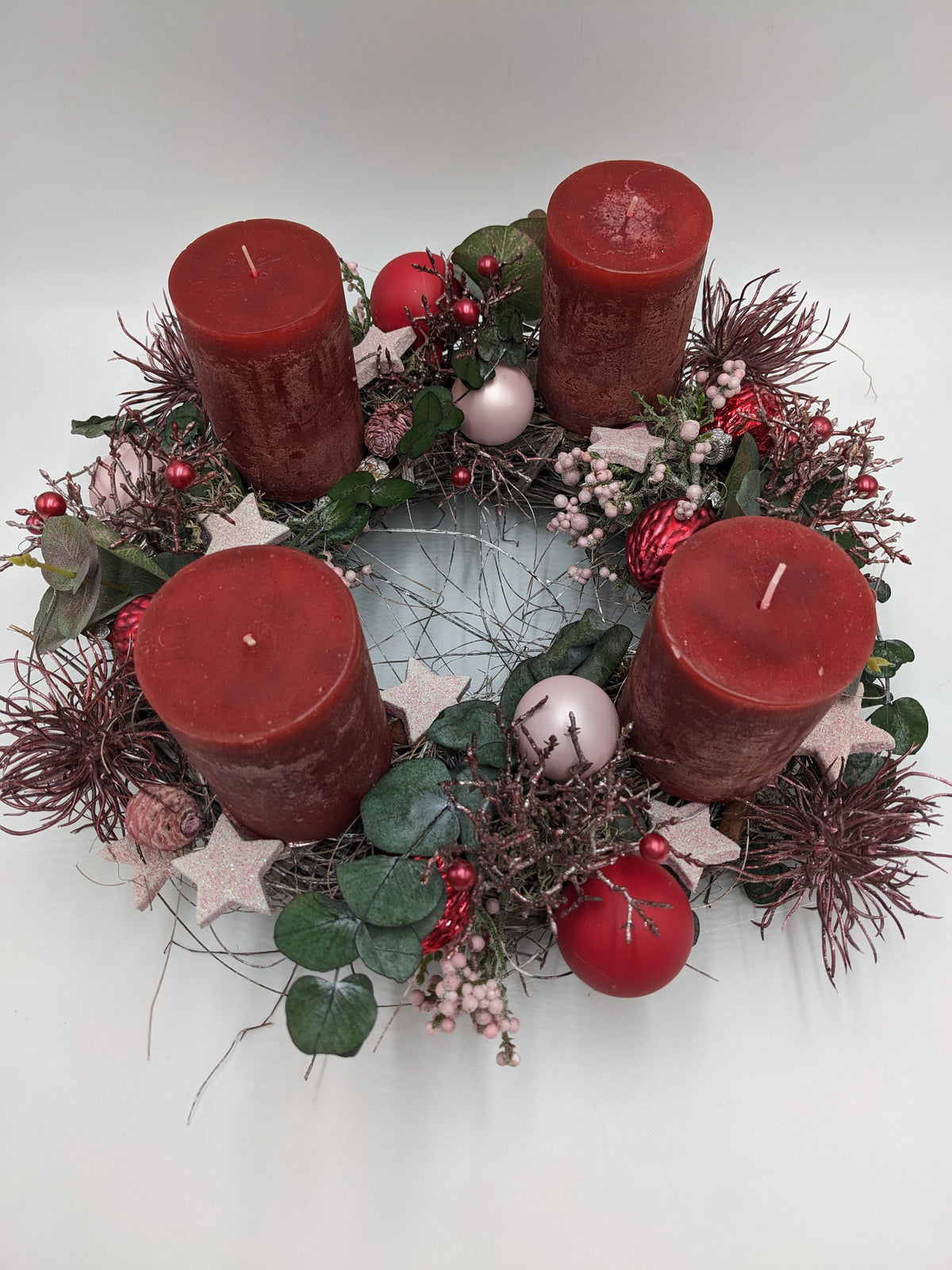 Adventsgesteck von oben: vier rote Kerzen im Kreis, rosa Glitzersterne, Beerenrispen, Zapfen und rötliche Spider-Blüten