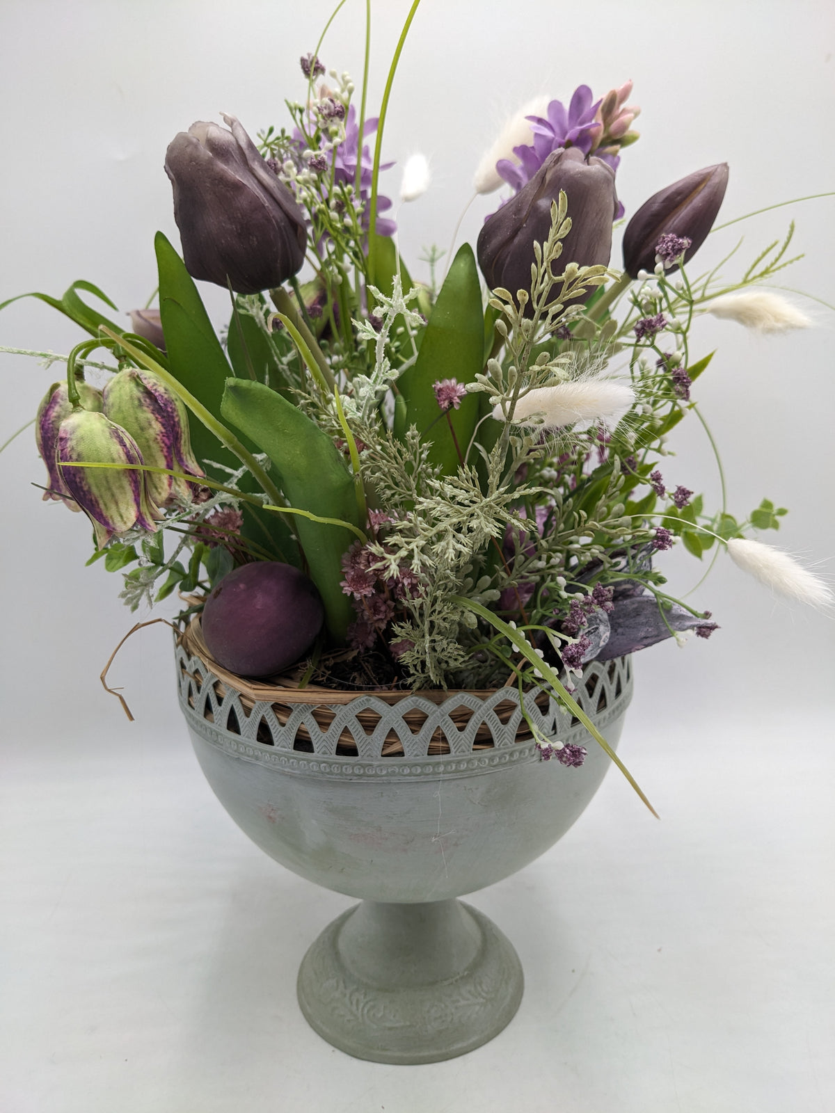Frühlingsgesteck mit dunkellila Tulpen, Schachbrettblumen und Hyazinthen in grauem Metallpokal, violettes Ei am Rand