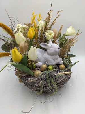 Osternest von vorne: Steinhase zwischen goldenen und grünen Eiern, dahinter gelbe Tulpen, Ähren und Zweige