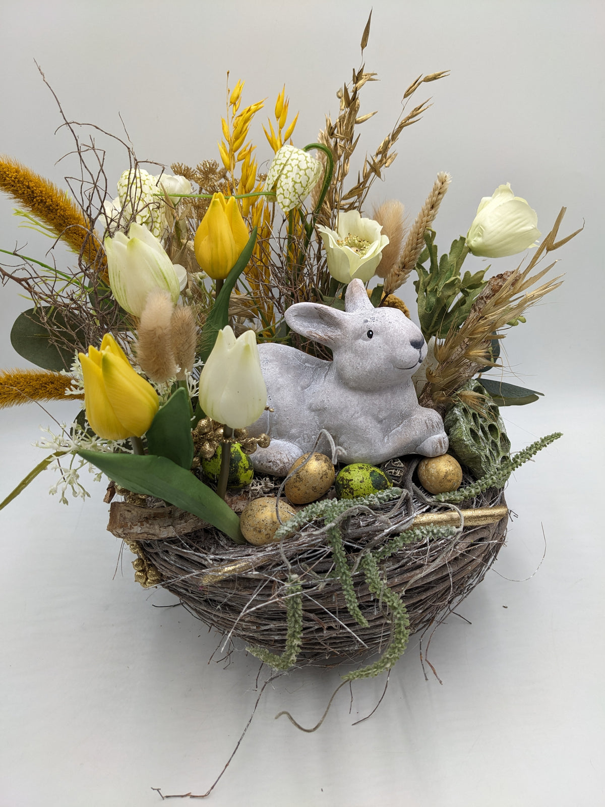 Osternest von vorne: Steinhase zwischen goldenen und grünen Eiern, dahinter gelbe Tulpen, Ähren und Zweige