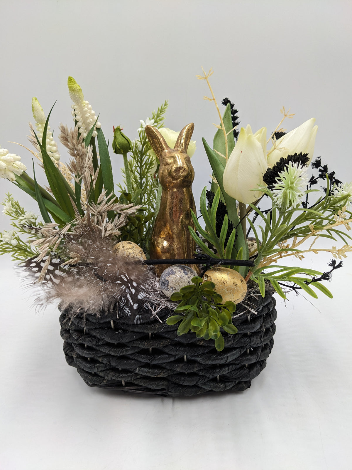 Ostergesteck im schwarzen Korb mit goldenem Hasen, weißen Tulpen, Traubenhyazinthen, Federn und gesprenkelten Eiern
