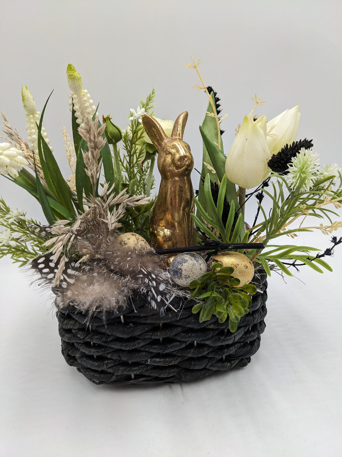 Ostergesteck von der Seite: schwarzer Flechtkorb mit Goldhase, cremeweißen Tulpen, Gräsern, Federn und goldenen Eiern