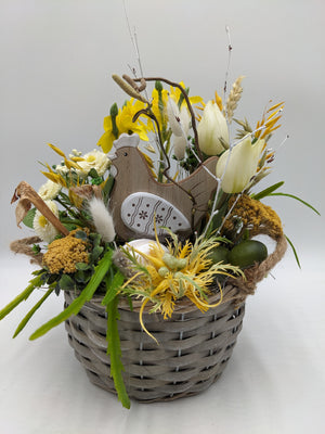 Ostergesteck im grauen Weidenkorb mit Holzhuhn, gelben Narzissen, cremeweißen Tulpen, Bellis und Ähren