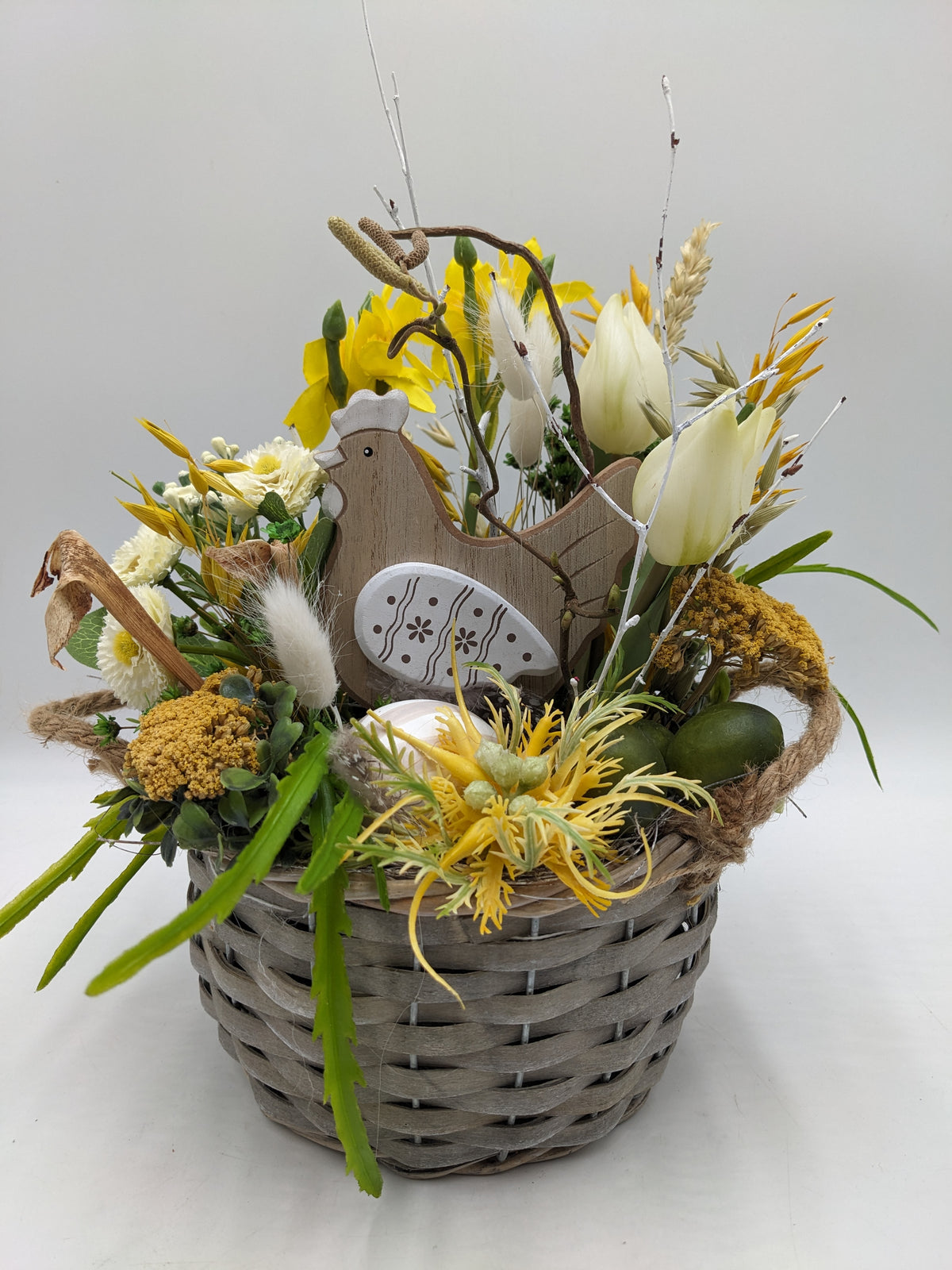 Ostergesteck im grauen Weidenkorb mit Holzhuhn, gelben Narzissen, cremeweißen Tulpen, Bellis und Ähren