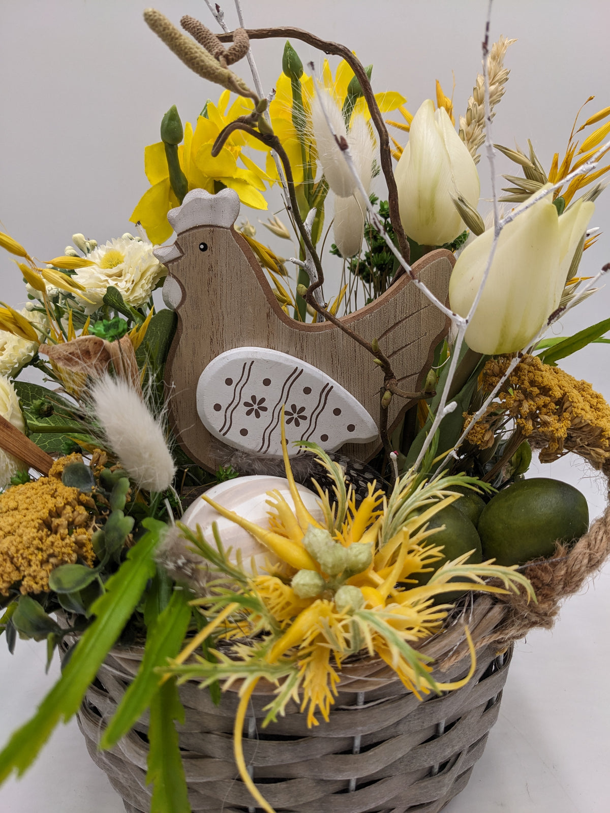 Nahaufnahme des Holzhuhns mit weißem Osterei, daneben grüne Eier, gelbe Schafgarbe und weiße Tulpen