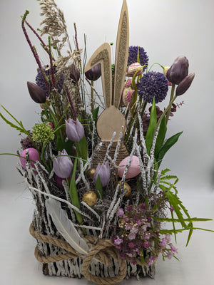 Ostergesteck im Geästkorb mit fliederfarbenen Tulpen, violettem Allium, Holzhase und rosa Eiern