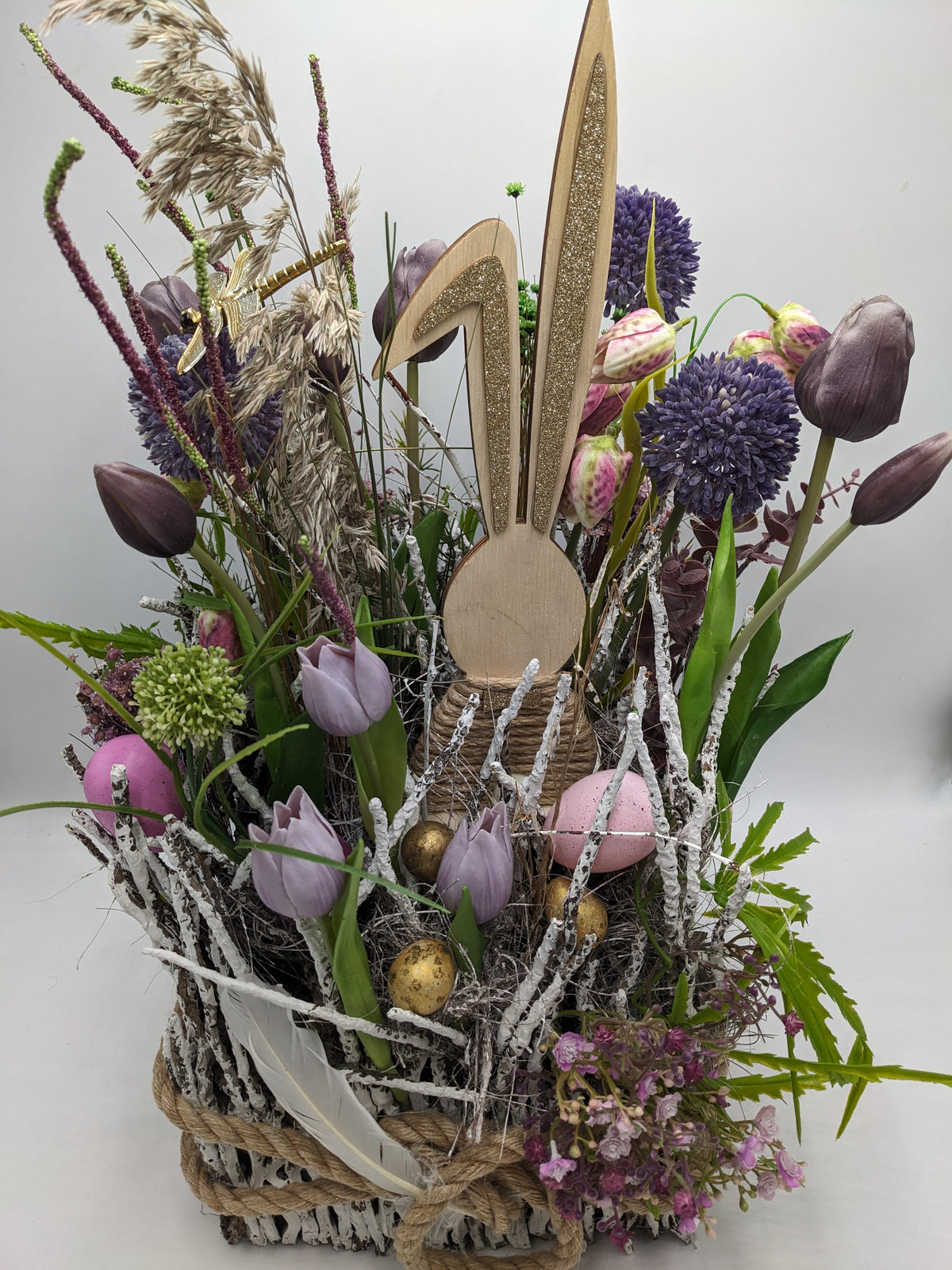 Ostergesteck von vorn: Tulpen, Schachbrettblumen, goldene Eier, weiße Feder und Gräser im Korb
