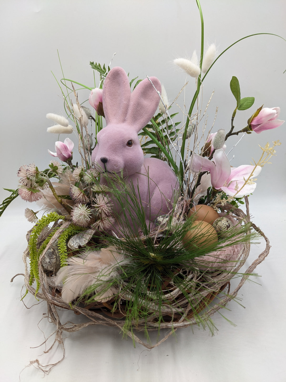 Ostergesteck im Zweigkranz mit rosa Hase, Magnolienblüten, Federn und braunen Eiern auf weißem Grund